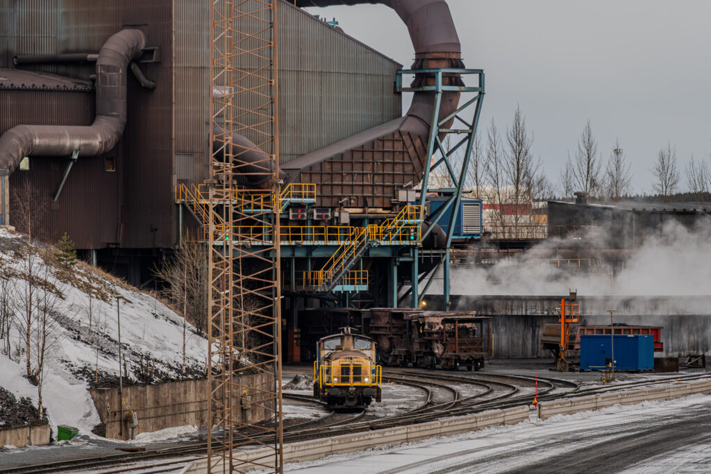 A photo of a train at the SSAB Luleå steelworks. The train is yellow. Two wagons carrying ladles can be seen in the background.